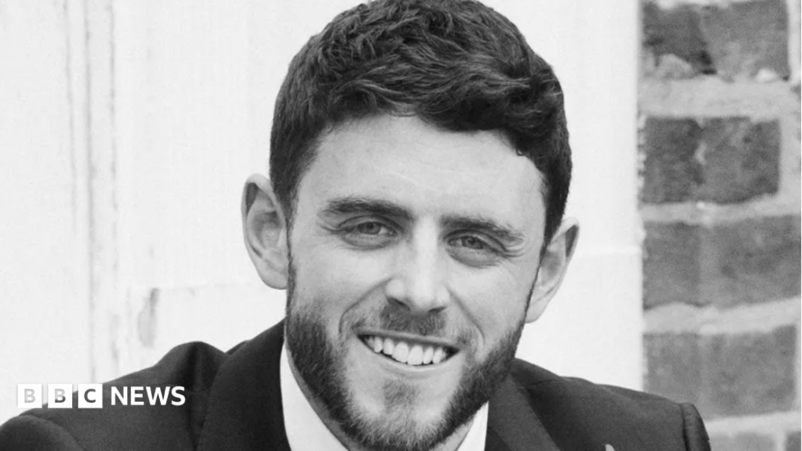 A black and white image of PC Andrew Harper a man with short brown hair and a slight beard wearing a suit and smiling at the