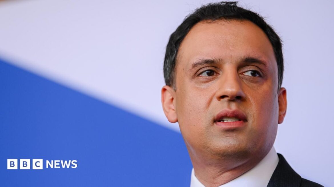 Anas Sawar stares into the distance. He has dark hair and is wearing a dark suit and white shirt. The background is blue and