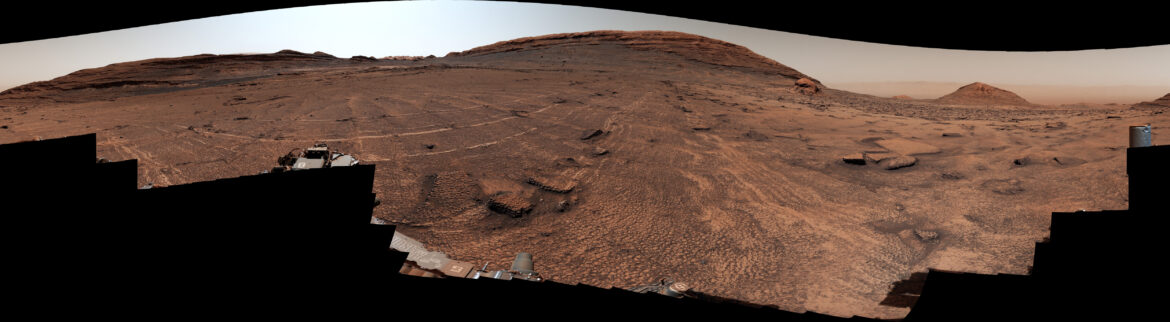 A panoramic view of a sweeping Martian plain covered in patterned fractures leading toward distant hills showcases the