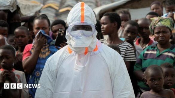 A health worker in personal protective equipment (PPE) stands near displaced people waiting for the burial of suspected