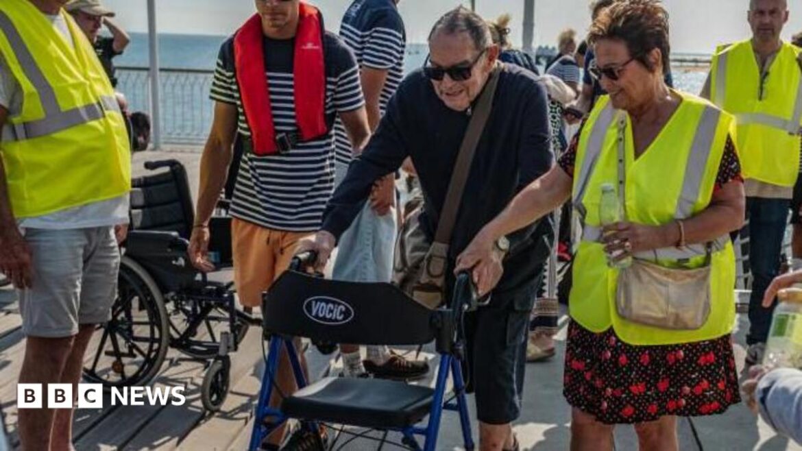 A man using a mobility aid walker is helped by volunteers dressed in yellow hi-vis jackets with the sea visible in the