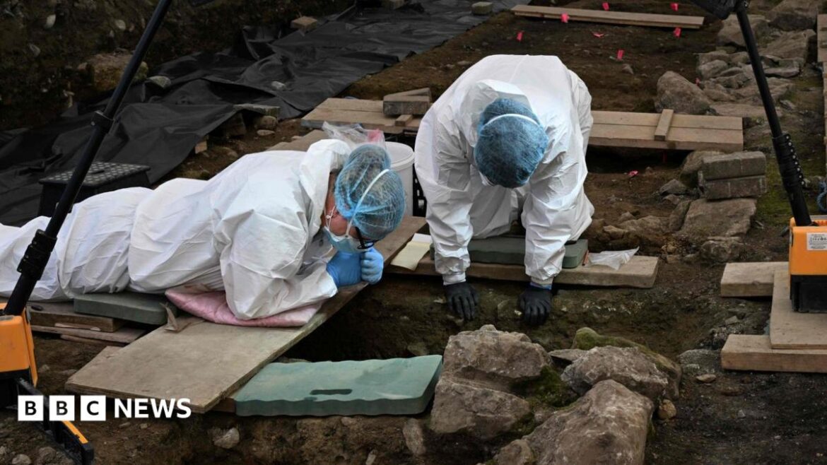 Two people in white excavation/forensic attire. They have blue nets over their hair and are wearing gloves. They are both