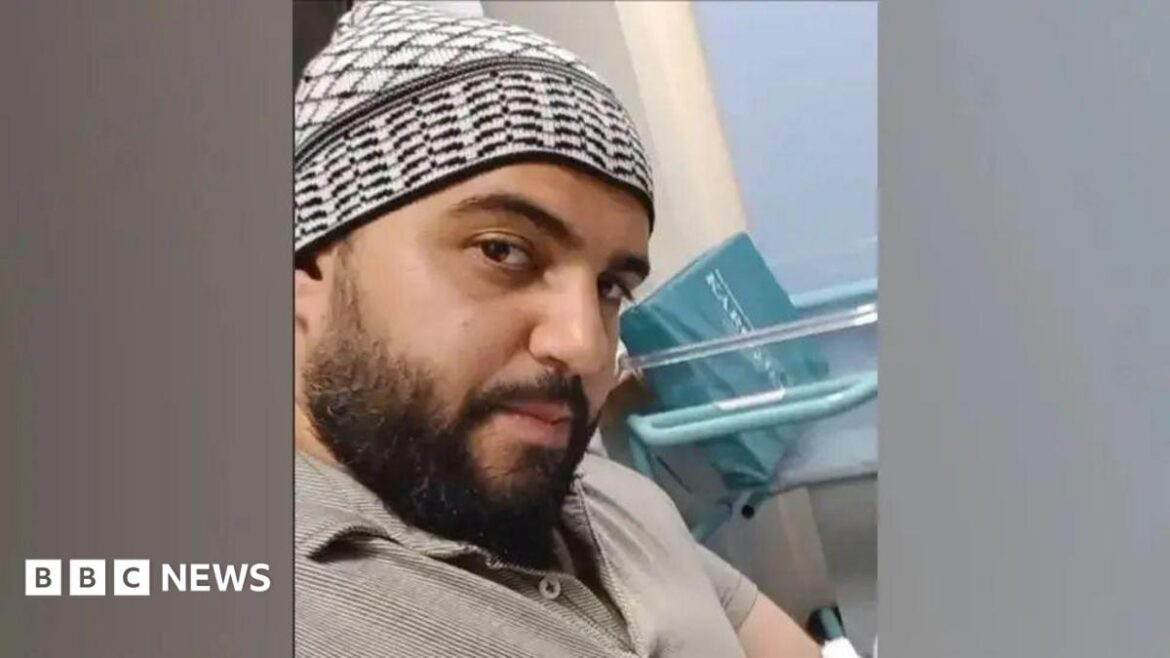 A selfie of a man who is sitting down in a hospital. He is looking directly at the camera. He is wearing a beige polo shirt