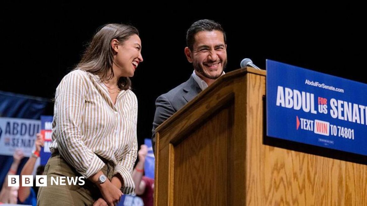 Alexandra Ocasio-Cortez smiles beside Abdul El-Sayed, who stands behind a podium with a sign that reads: Abdul for US Senate