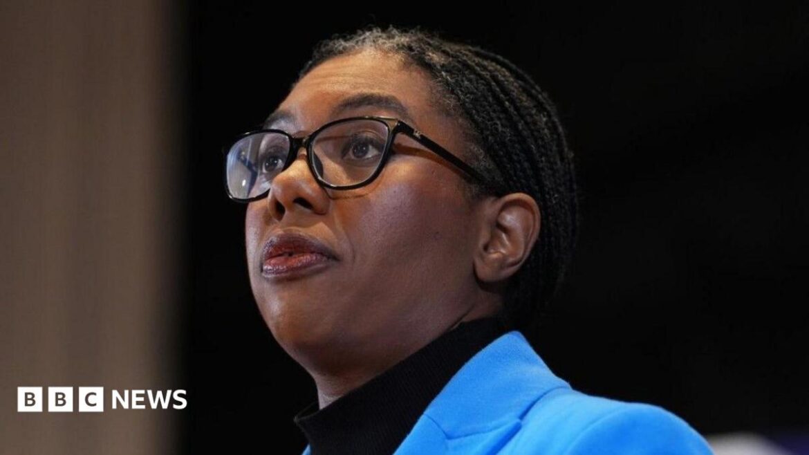 Kemi Badenoch has a serious expression and is wearing a sky blue jacket over a black turtleneck top. She has her braided