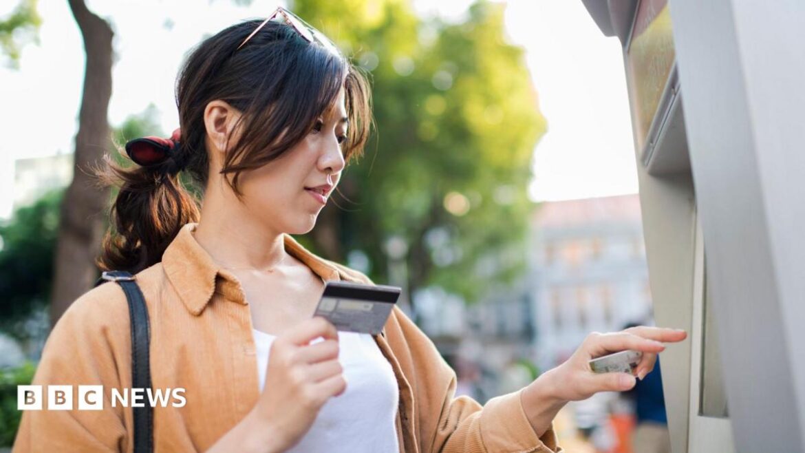 A woman uses a cash machine on the street on a sunny spring day. She holds a credit card in her hand and is pressing buttons