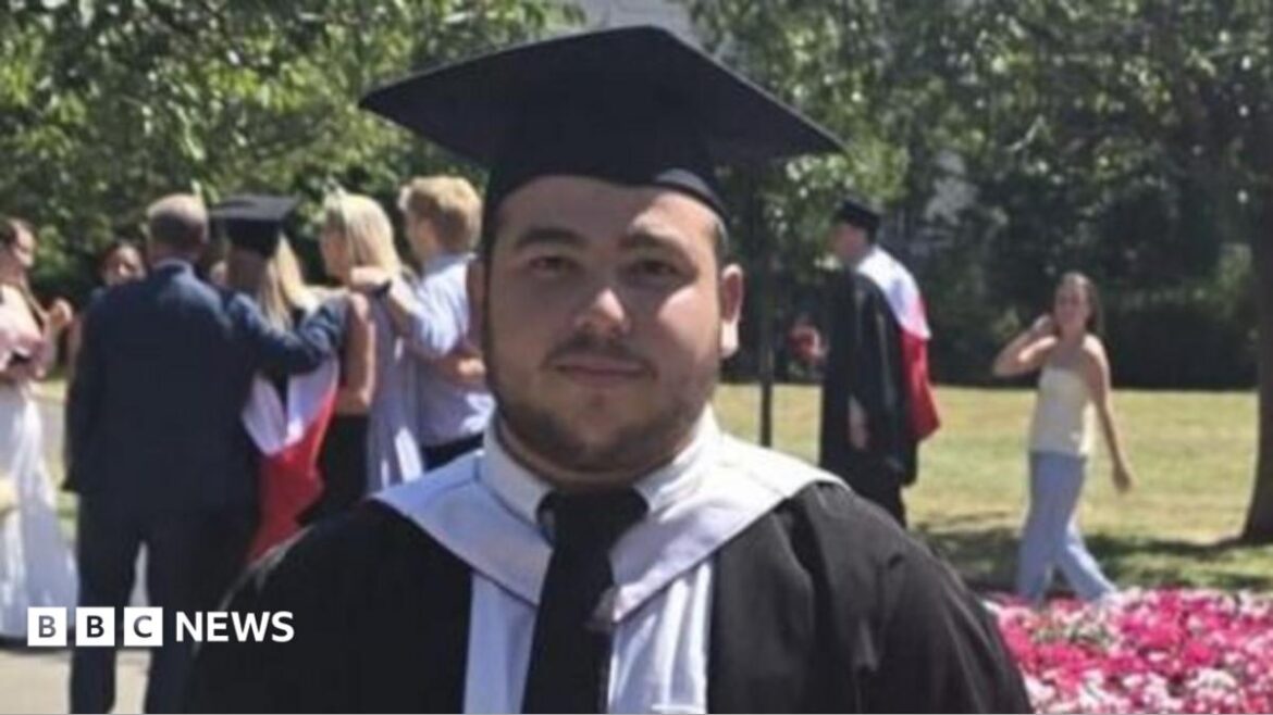 Oliver, who has dark hair and short facial hair, is wearing graduation robes and a cap, standing beside a flower bed in a