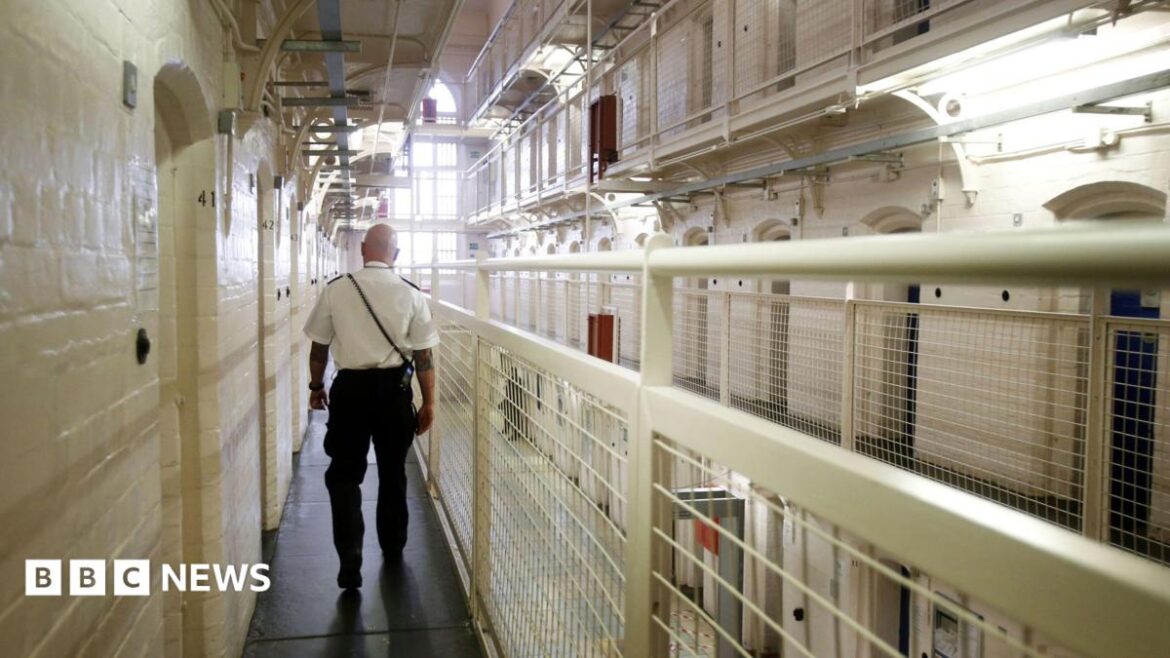 Prison Guard stands in a prison with his back to the camera he is wearing a white shirt and black trousers