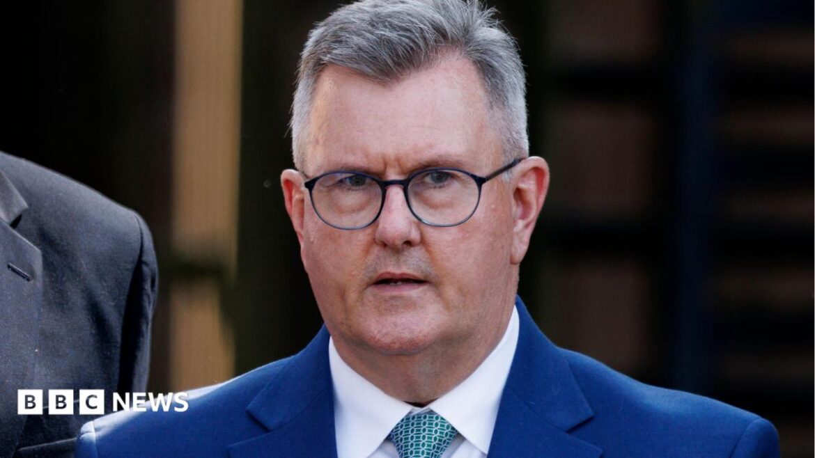 Jeffrey Donaldson has short grey hair, black round-rimmed glasses and is wearing a navy suit jacket over a white shirt and