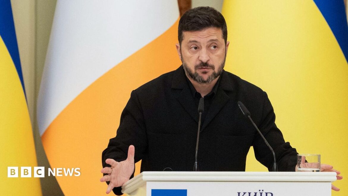 Zelensky looks to the right of frame while gesturing with his hands as he speaks from behind a lecturn with the Ukrainian