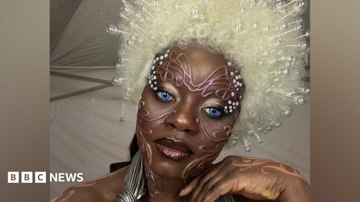 A person in bold avant‑garde makeup with bright blue eyes, a large white beaded wig, and metallic costume accessories