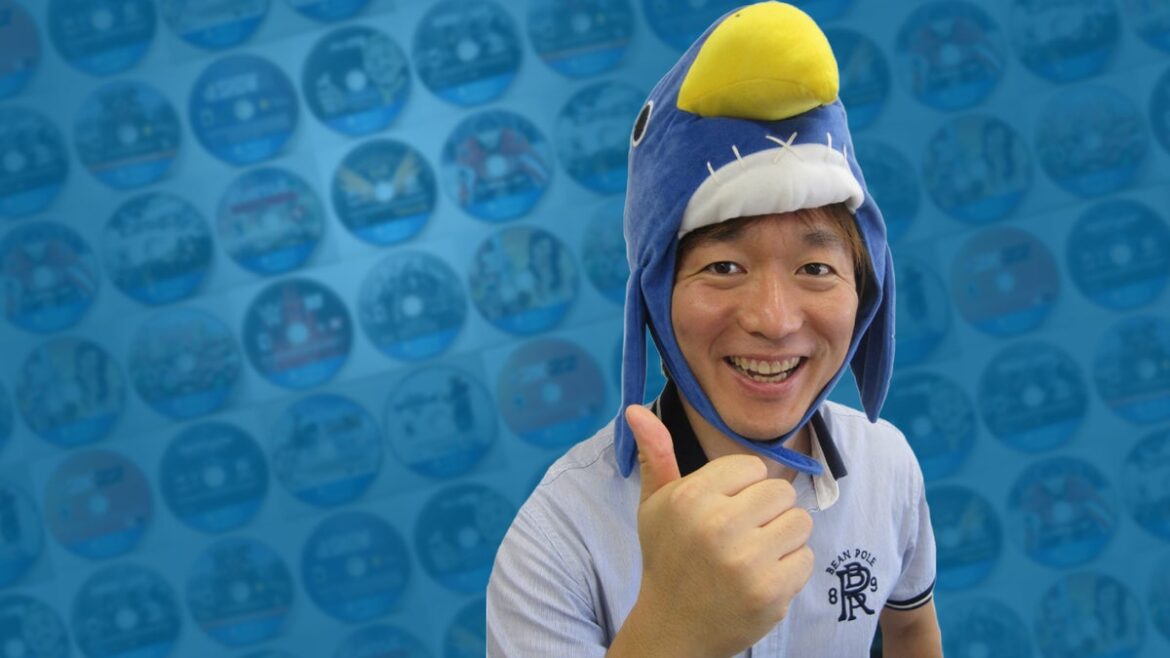Sohei Niikawa in a prinny hat, in front of a grid of PS4 discs