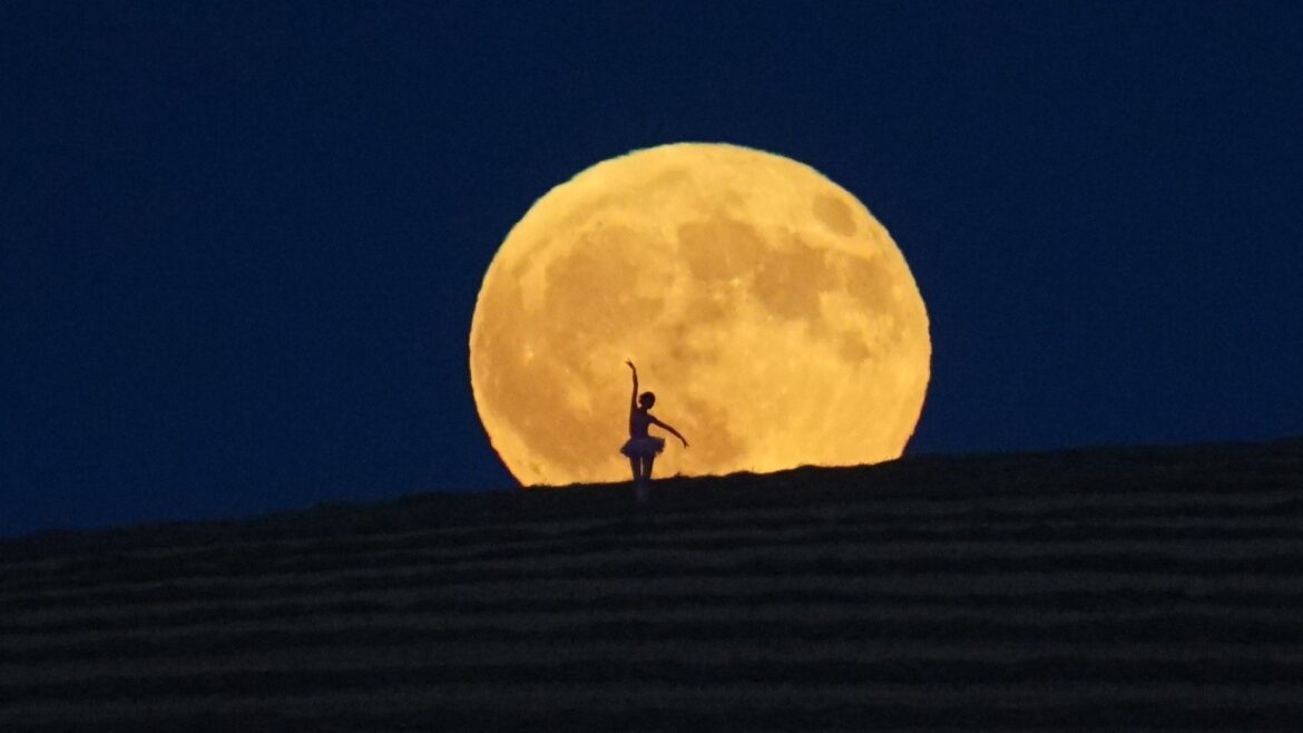 The silhouette of a ballerina is shown dancing in front of the disk of a full moon as it rises over a hillside in a dark