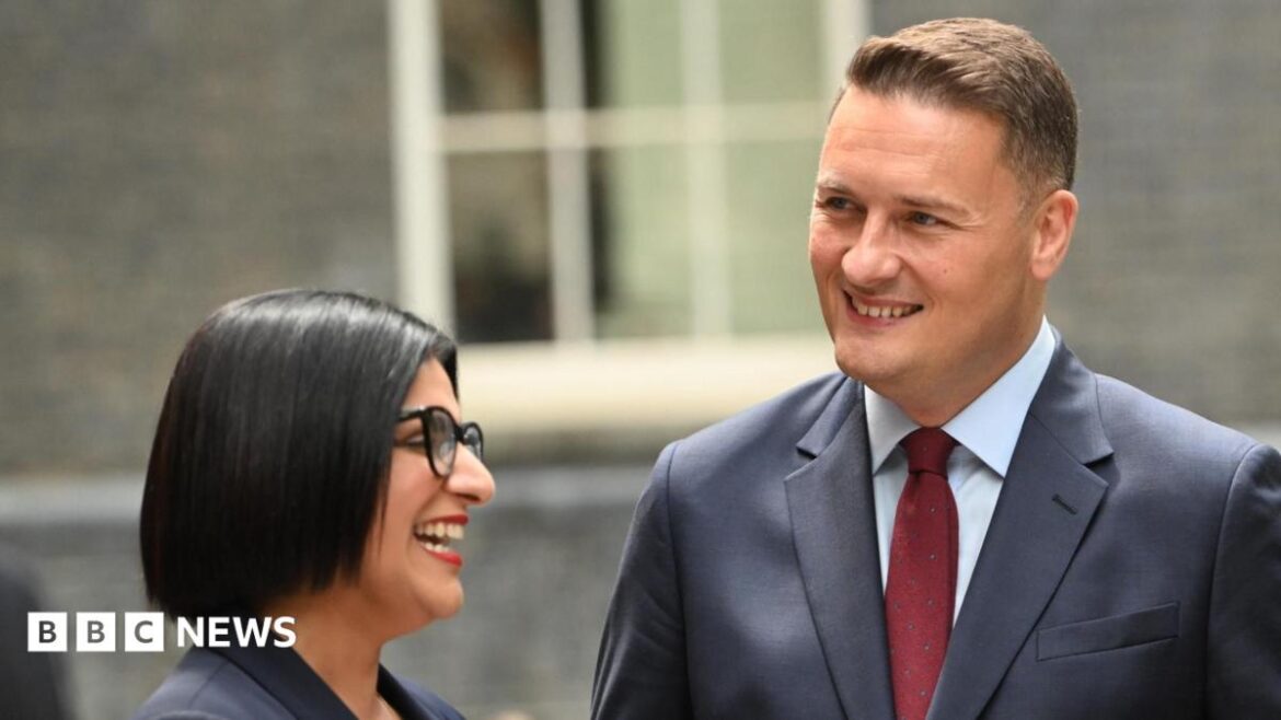 Home Secretary Shabana Mahmood wears a dark suit jacket, red lipstick and black glasses as she laughs with Defence Secretary