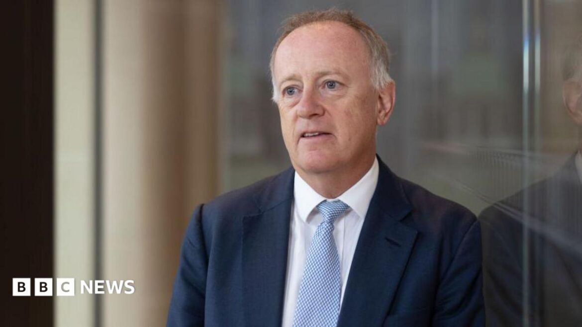 John Neal, former chief executive officer of Lloyd's of London, leaning against a glass wall in an atrium outdoors. He is