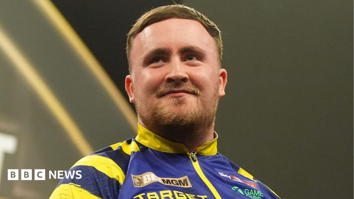 Luke Littler, pictured in March 2026, wearing a blue and yellow jacket during a darts tournament