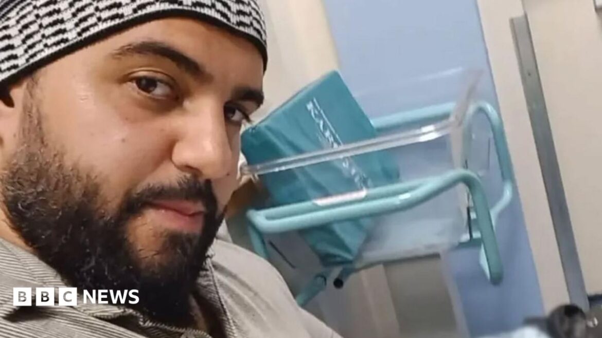 A selfie of a man who is sitting down in a hospital. He is looking directly at the camera. He is wearing a beige polo shirt