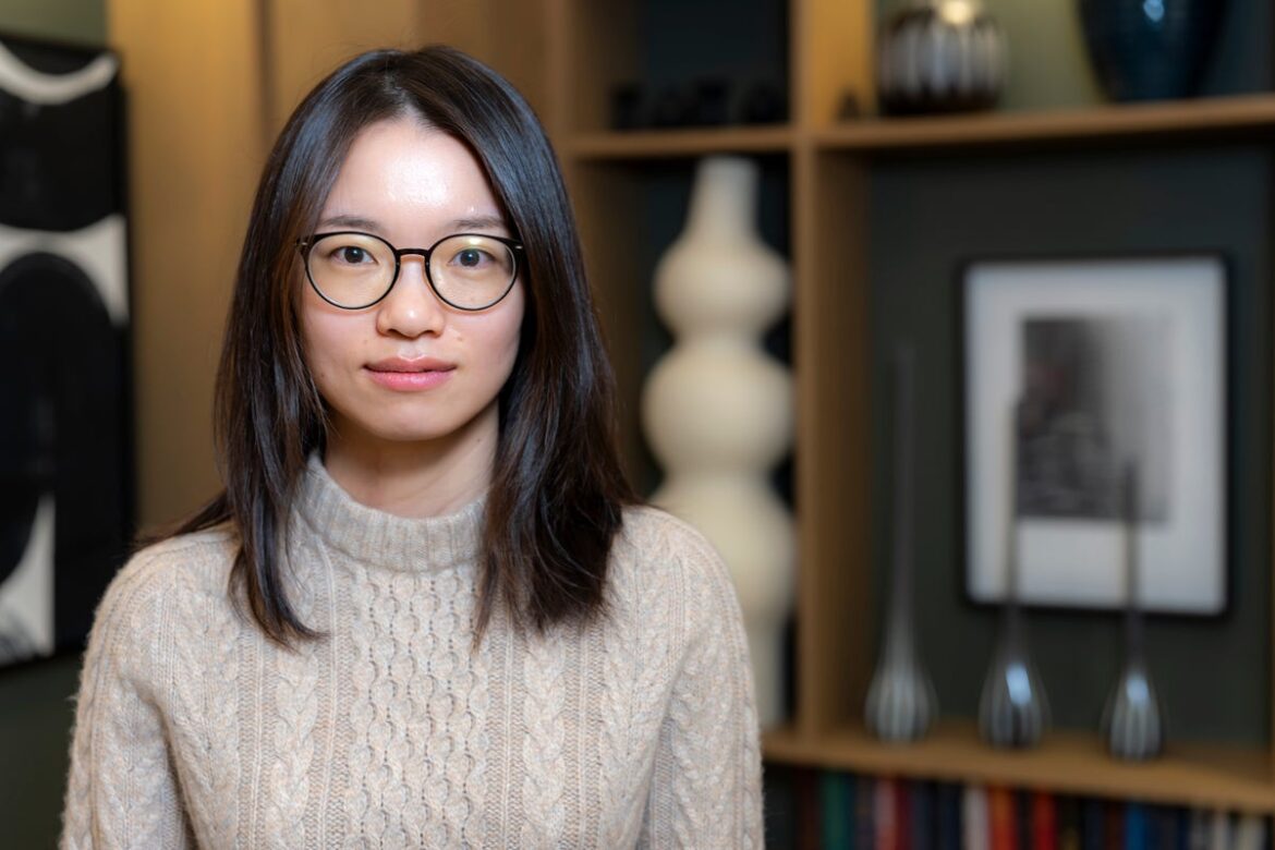 A woman in glasses stands against an out-of-focus bookshelf
