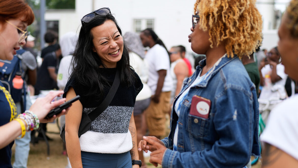 Francesca Hong smiles while standing and facing an out-of-focus person in the foreground who is speaking to her, with to