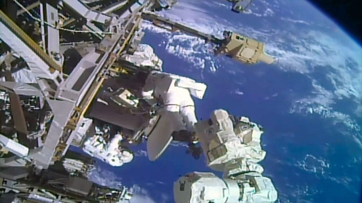 two astronauts in white spacesuits work near a large antenna dish outside a space station with a blue and white planet as