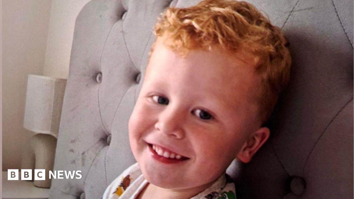 Three-year-old Noah Woods, with light hair and wearing a brightly coloured top, smiles at the camera