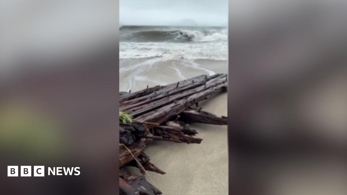 A wooden shipwreck washed up on a beach with big waves in the background