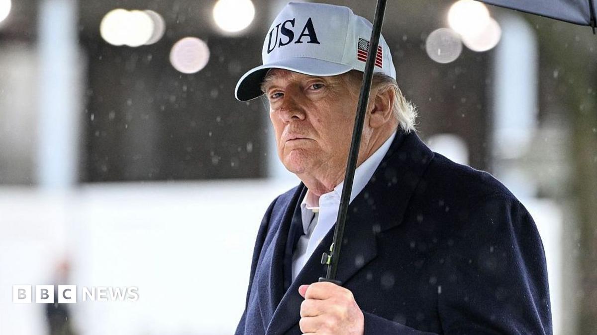 President Trump holds an umbrella as rain drops fall around him. He wears a dark suit and a white baseball cap with USA