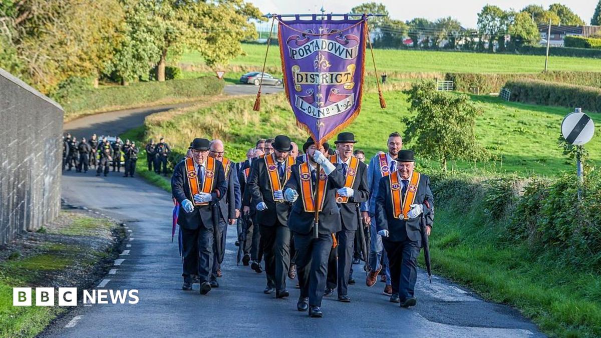 Orangemen walk down a road, behind them is a police presence