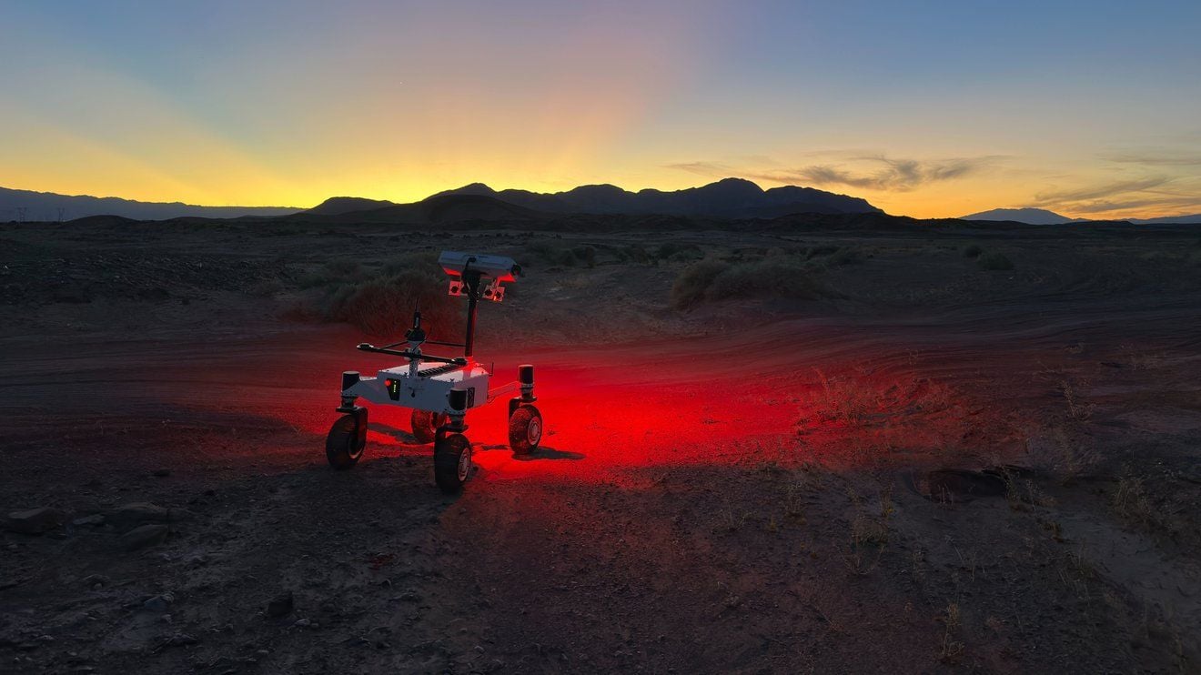 a small, four-wheeled rover shines a red light on a dark desert ground and the sun sets behind silhouetted hills in the