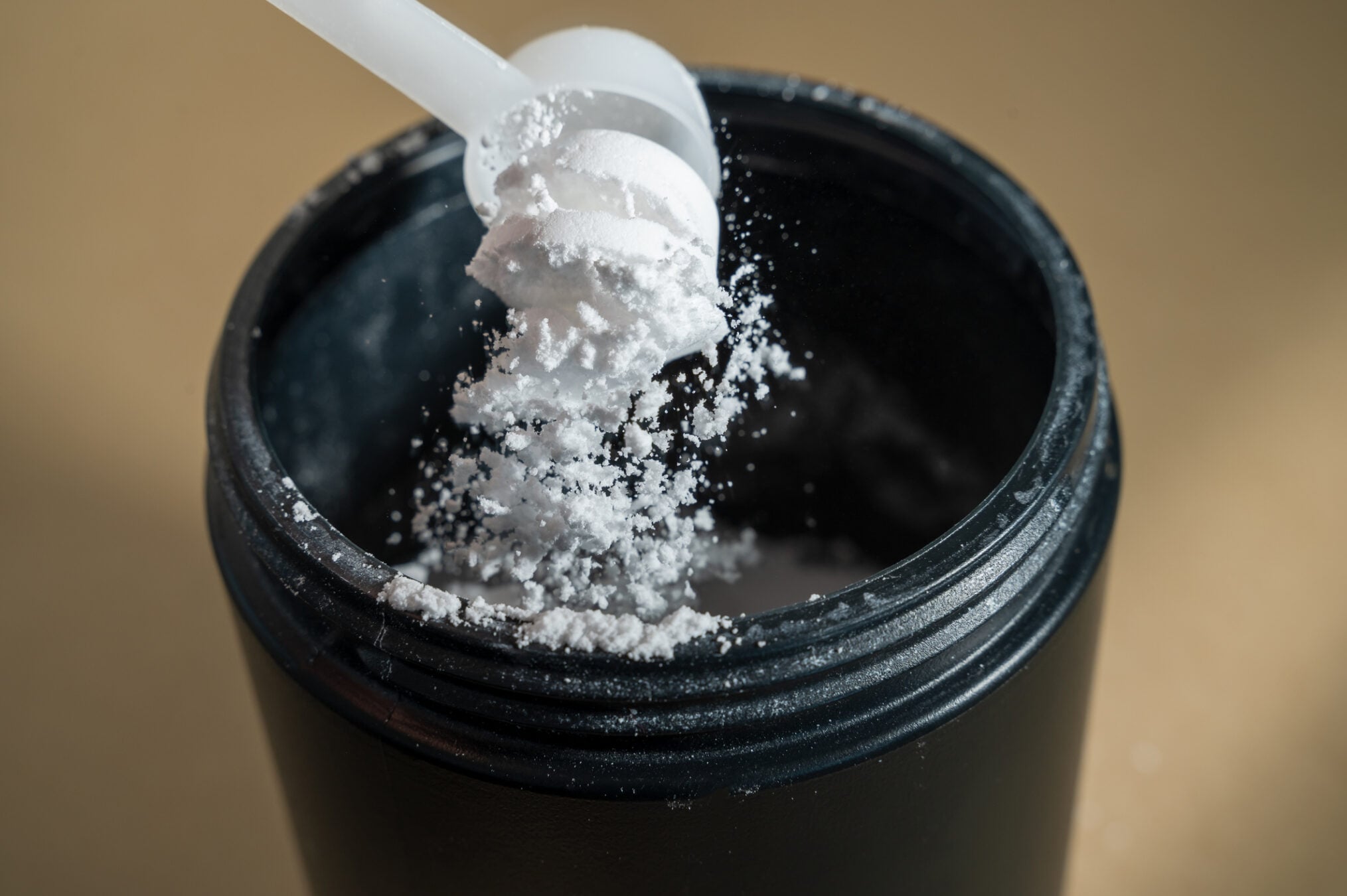 Creatine powder pouring from measuring scoop into jar on beige background
