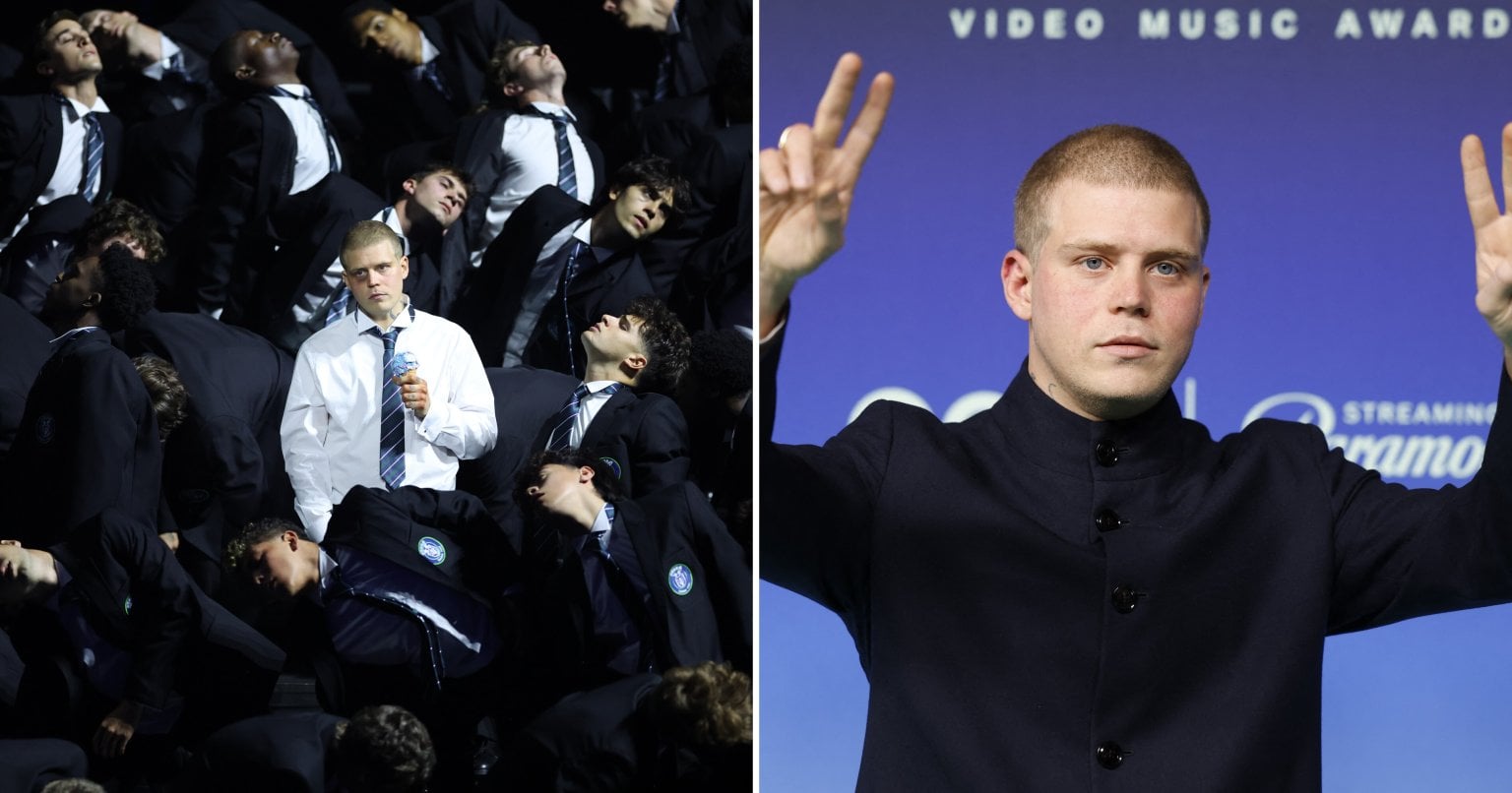 A young man in a white shirt and tie holds a blue flower and stands still amid a crowd of suit-clad dancers leaning back