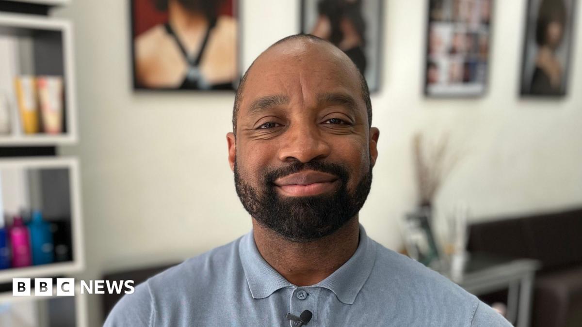 Marcus is sitting on a chair in his hair and beauty salon. He is black, with short hair and a moustache and beard. He is