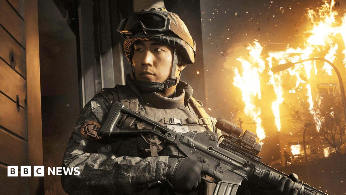 An in-game screenshot showing a male Korean soldier holding a gun with a building on fire behind him