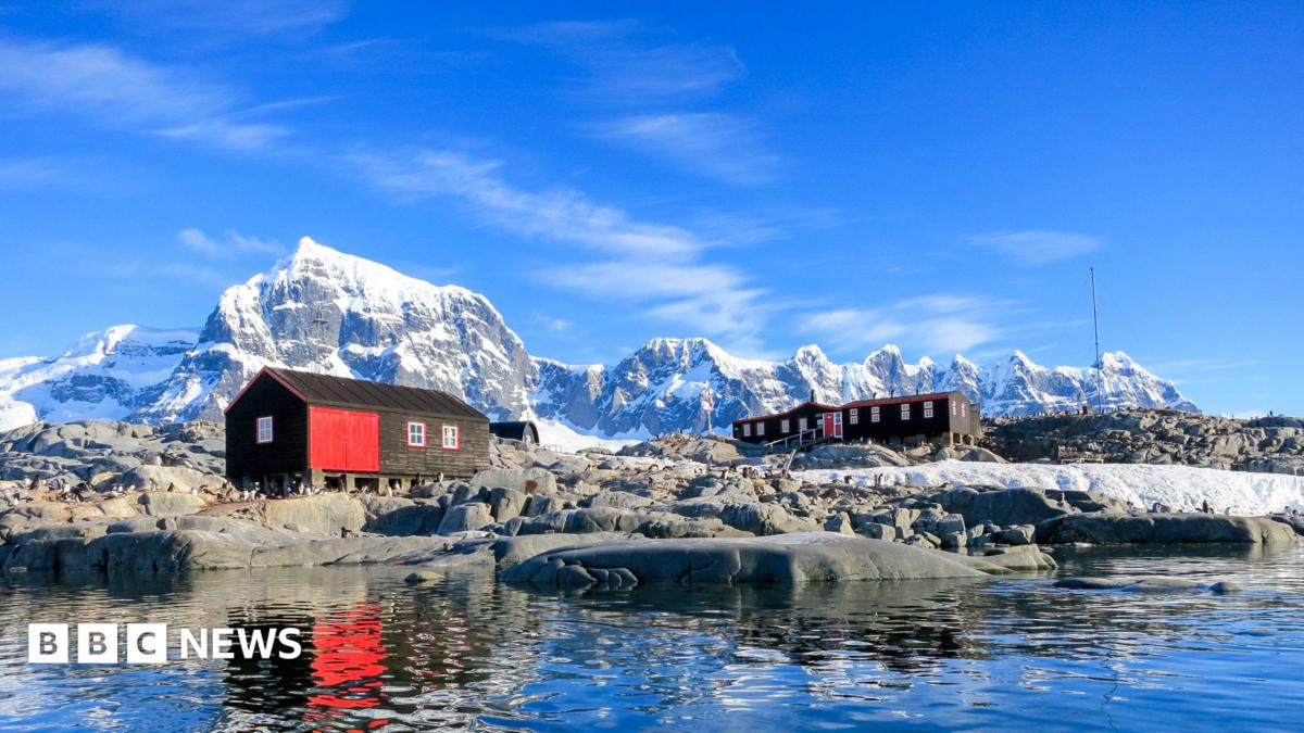 Two red and black huts sit on rocky ground beside calm Antarctic waters, with snow-covered peaks rising in the background