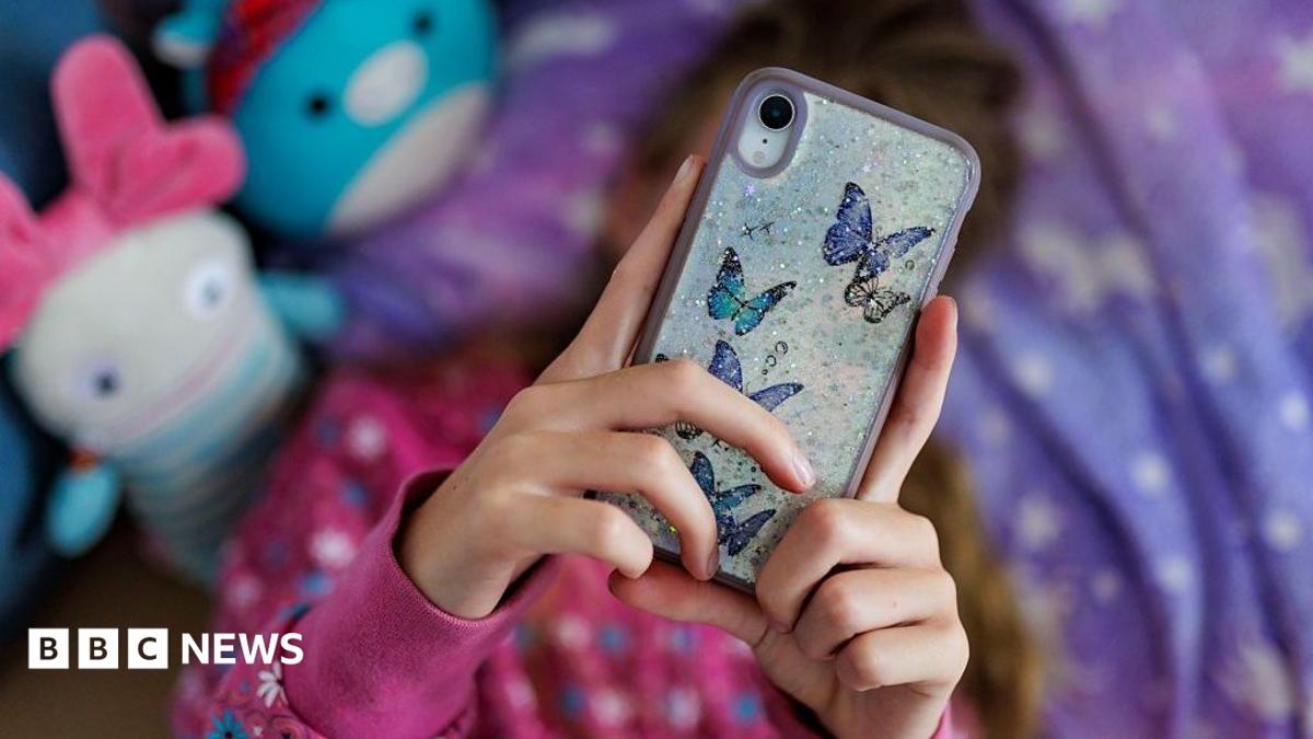 A young girl with long brown hair lying on her bed with a purple and white duvet, looking at her phone over her face, which
