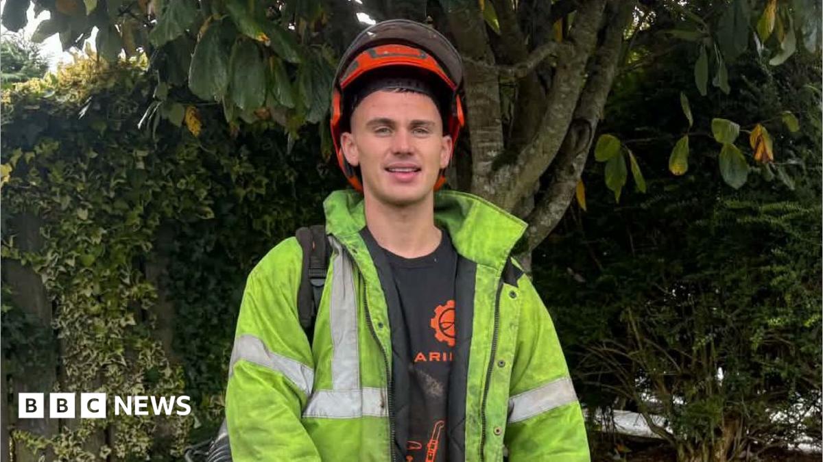 Callum Kirk is wearing a orange helmet, a yellow his-vis jacket, with a black t-shirt underneath. He has equipment in his
