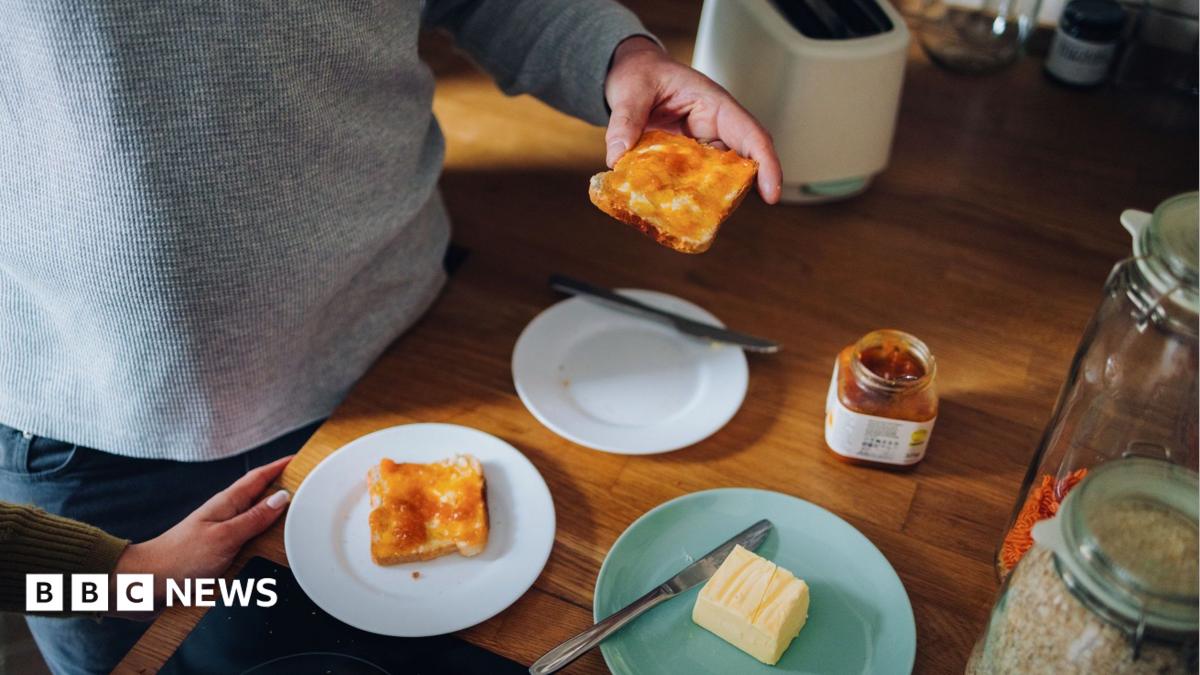 A man is visible from the chest down standing at a table eating marmalade on toast with a woman's hand visible in the shot