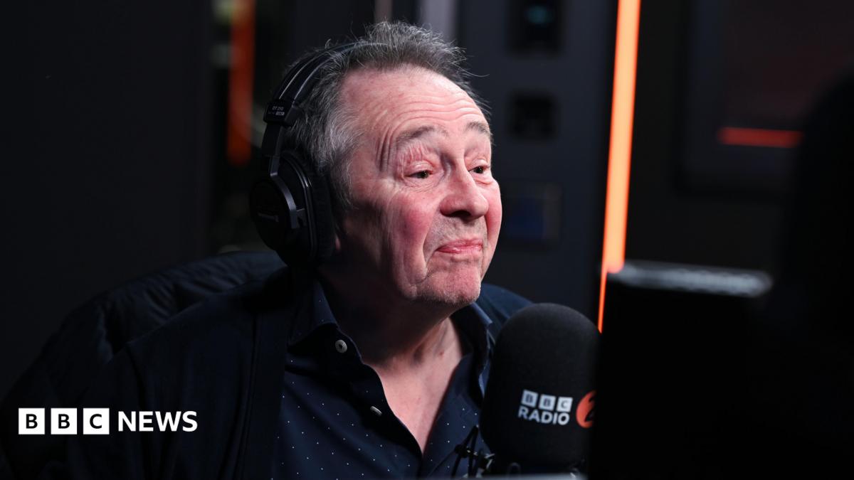 A man with grey hair and wearing a large set of headphones sits behind a BBC Radio 2 microphone