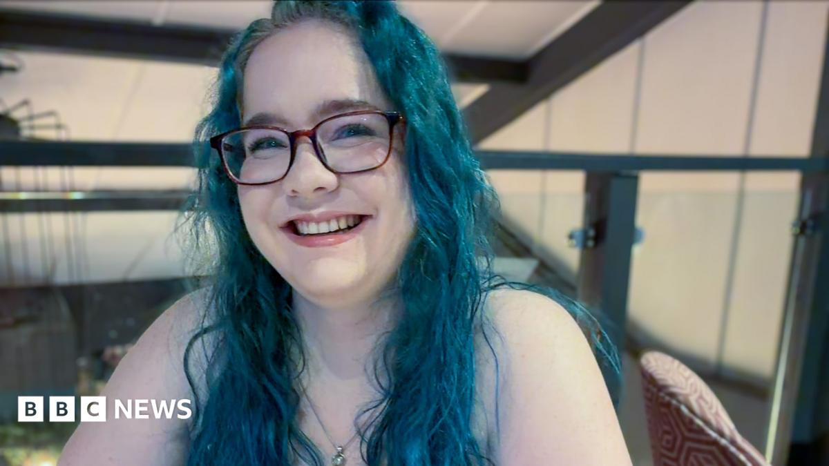 Olivia Miller-Davis, with long blue hair and glasses, sits smiling in a bar