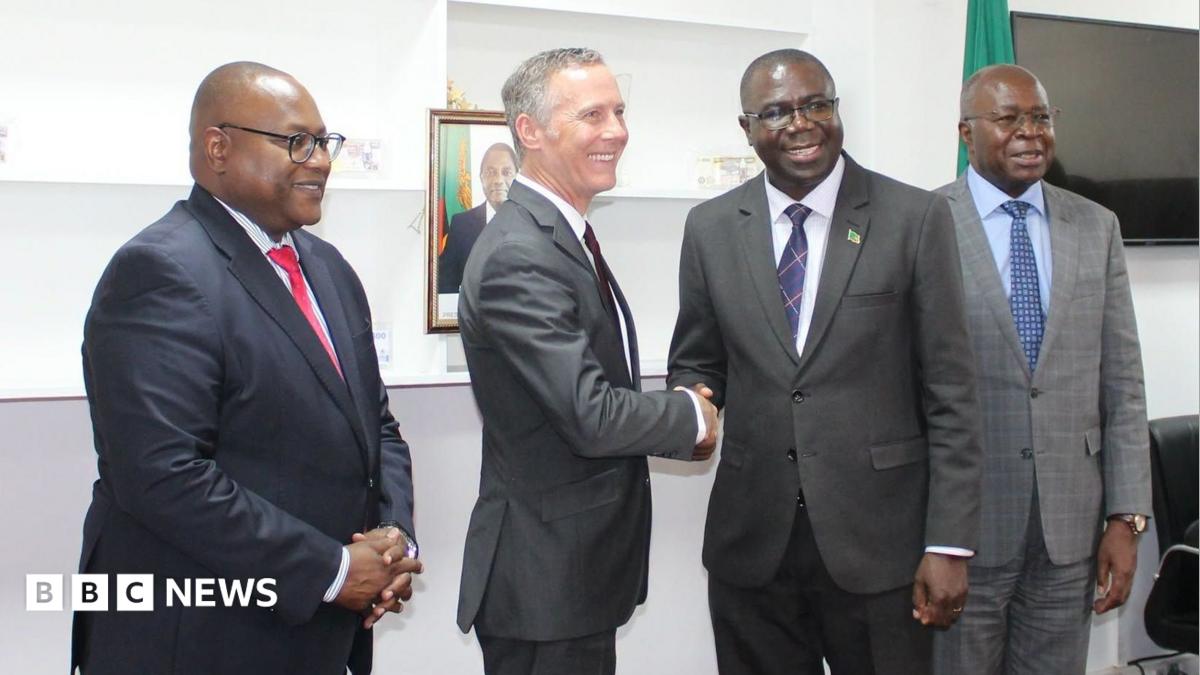 Four men stand in suits, two shaking hands and smiling in the middle