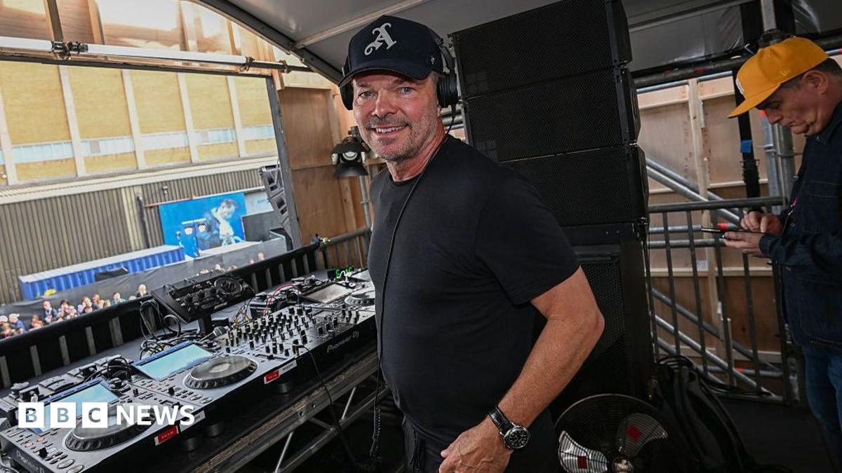 Pete Tong stands beside DJ equipment on a stage platform wearing headphones and a dark T-shirt. Mixing decks, speakers and