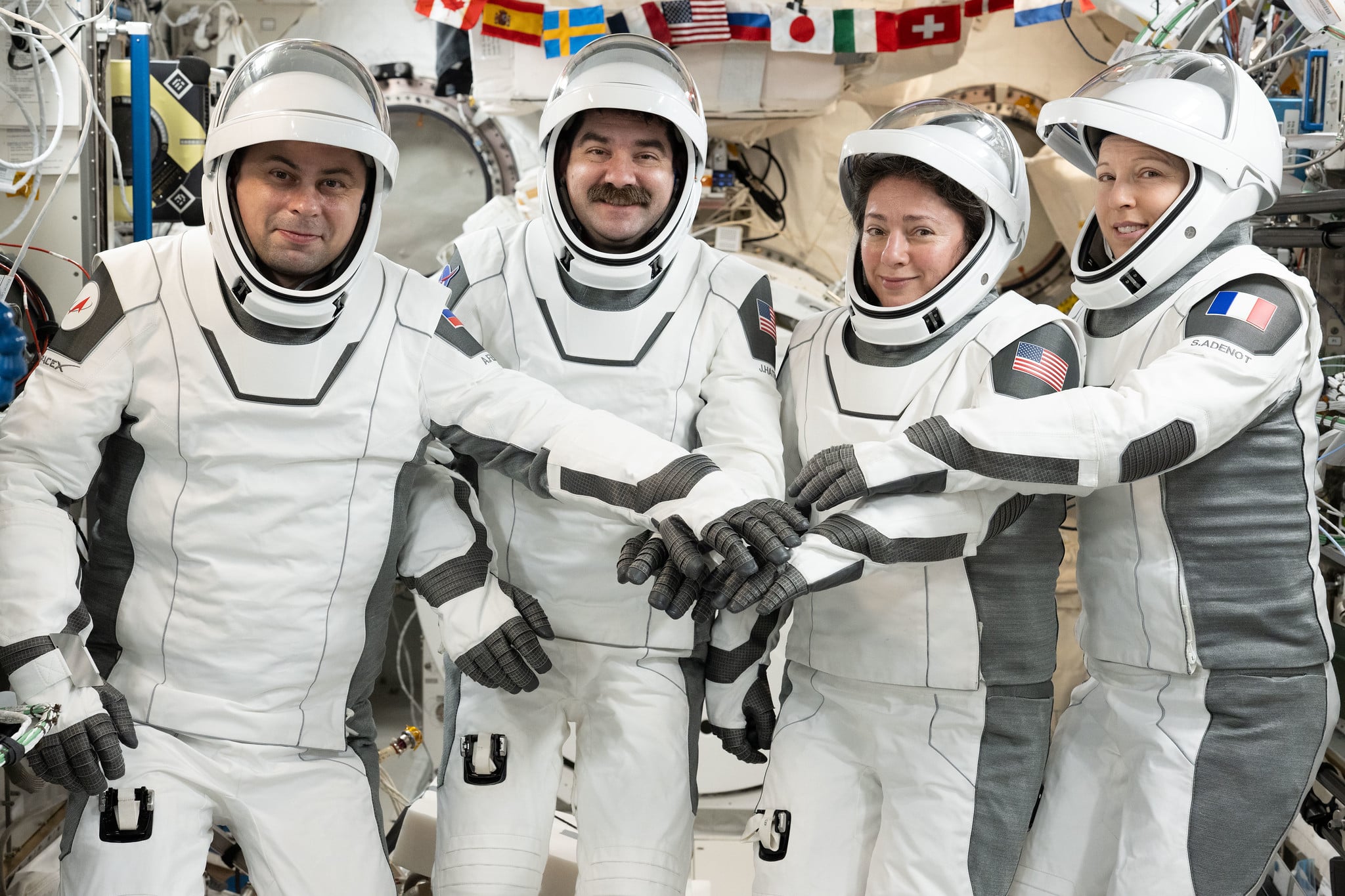 Four astronauts in white space suits smile at the camera and put their gloved hands together aboard the space station