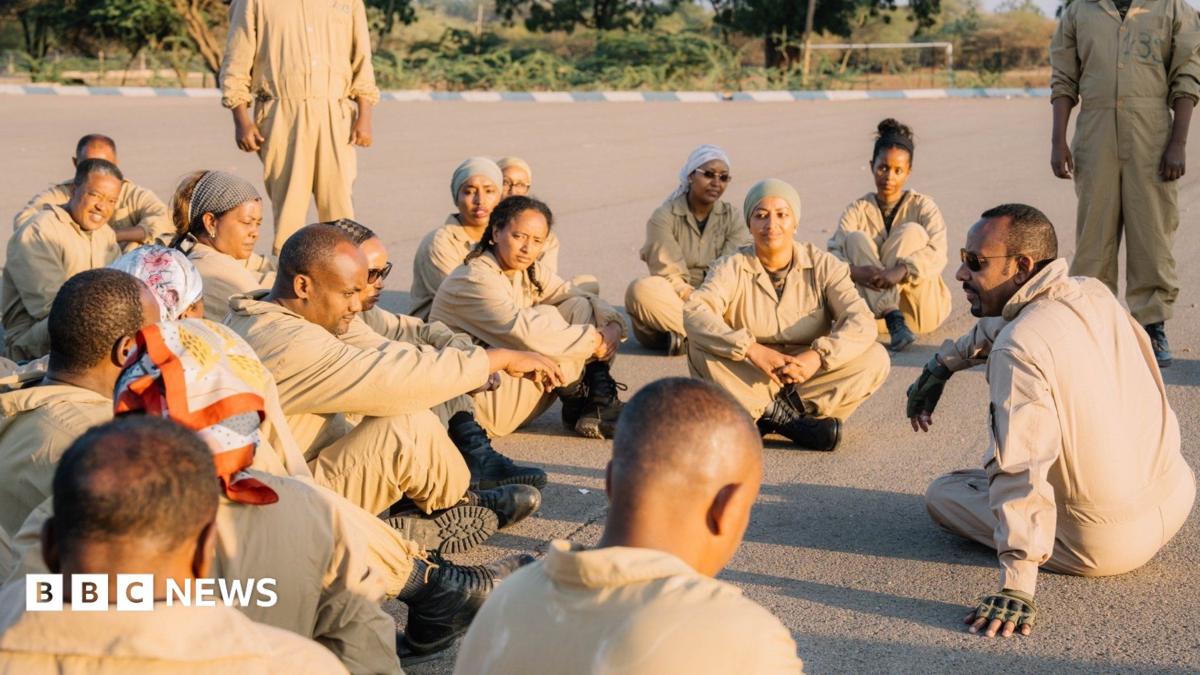 A photo of Abiy Ahmed seated on the ground, while wearing brown military attire. He is speaking to a group of people wearing
