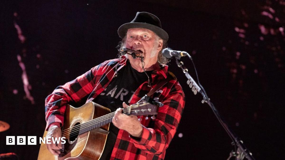 Neil young wears a red and black checked shirt and plays a guitar