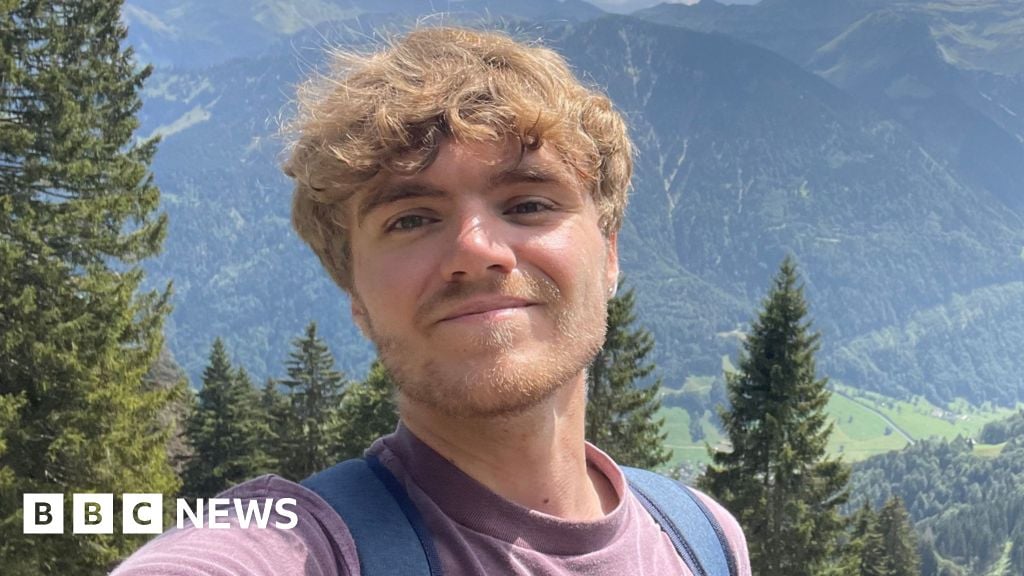 A selfie style picture of Tom standing in a lush green landscape with mountains in the background. The 24-year-old wears a