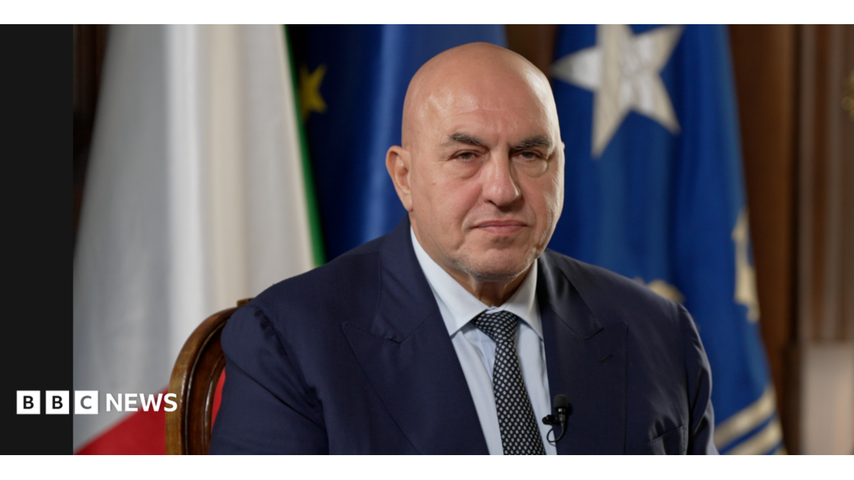 Guido Crosetto poses wearing a suit in front of the Italian and EU flags