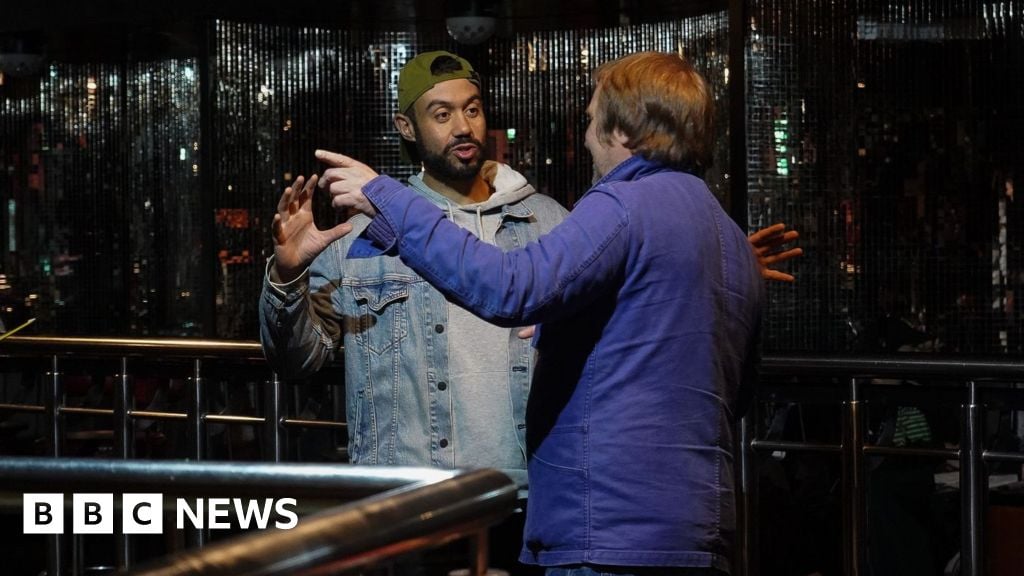 Two men are standing in a nightclub and appear to be talking. There is a shimmery backdrop on the walls