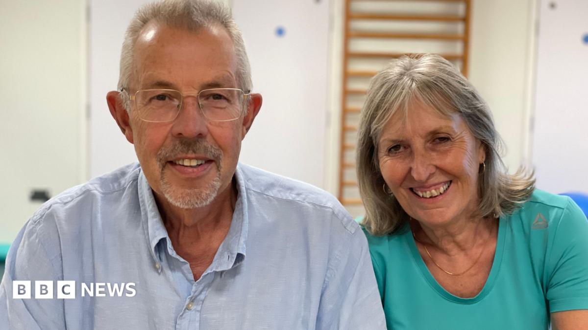 Steve and Angela Bucksey sit side-by-side smiling - Steve has grey hair and is wearing a light blue shirt with a collar, and