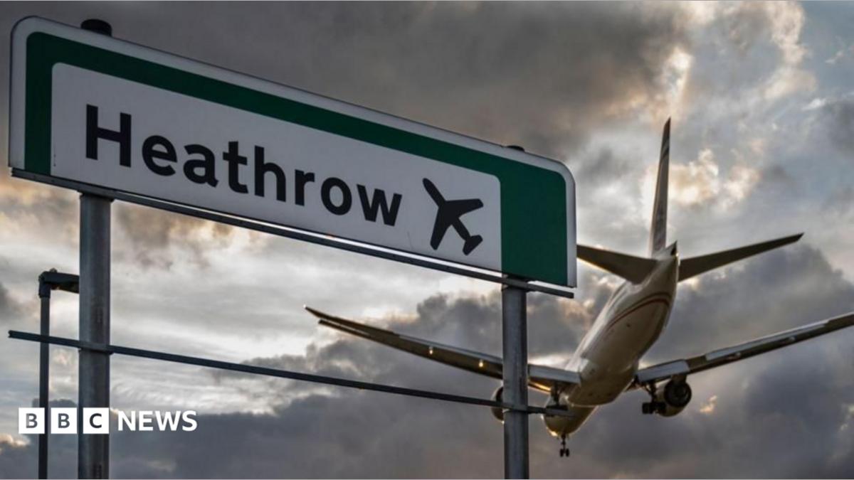 A plane flies by a road sign saying Heathrow on it. It's a cloudy, gloomy sky