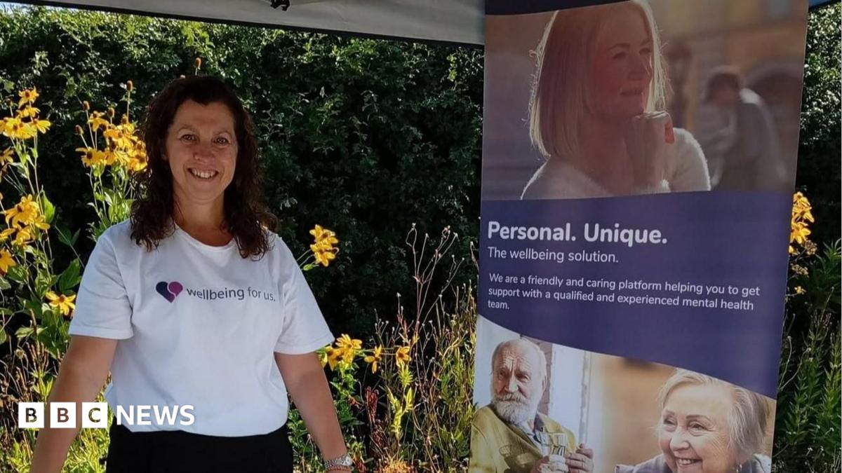 A woman with dark hair and wearing a white t-shirt with a wellbeing logo stands next to a display stand featuring several